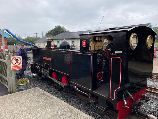 A black, boxy steam engine with a driver filling it up using a blue hose. The driver is standing next to the train, close to their full height, and they're about as tall as the train.